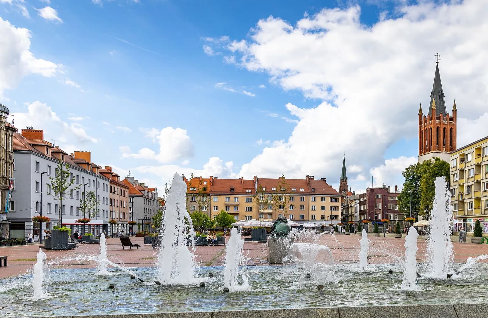 A town square with active fountains in the foreground and historic buildings with a church steeple in the background under a partly cloudy sky.